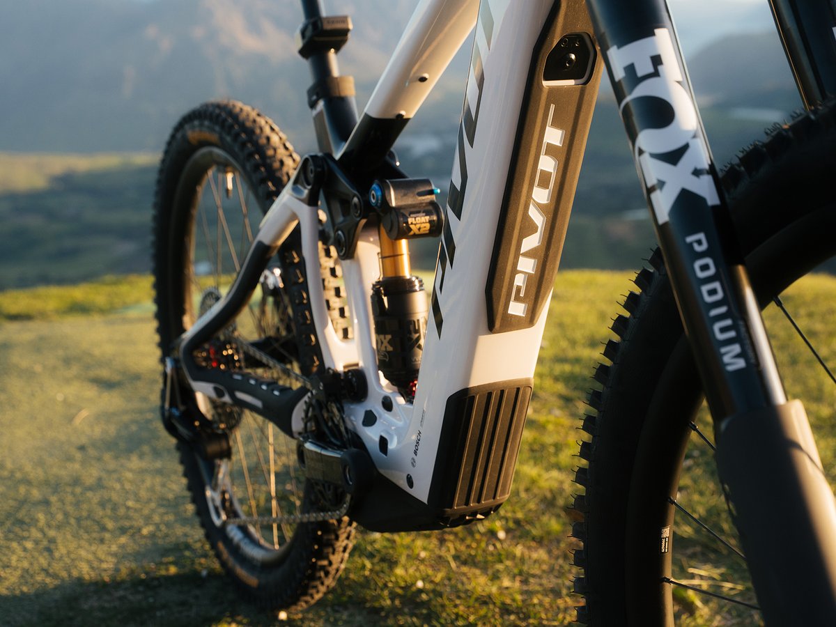 A close-up of a white Pivot mountain bike with Fox Podium suspension shown on a grassy hillside at sunset, focusing on the frame, front and back suspension, and front tire. 