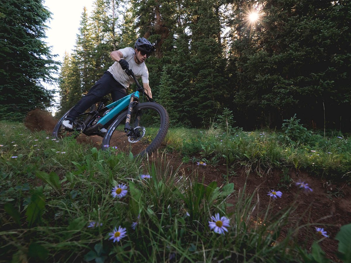 Mountain biker railing a bermed dirt corner on a teal Yeti full-suspension bike, sun flaring through spruce trees and wildflowers.