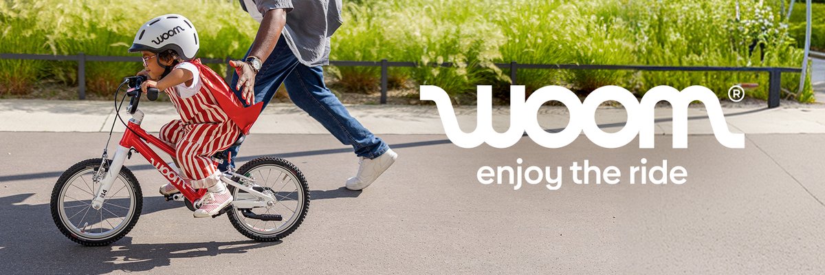 A child in a red striped outfit learning to ride a red woom bike, steadied by an adult running alongside them on a sunny path. woom — enjoy the ride.