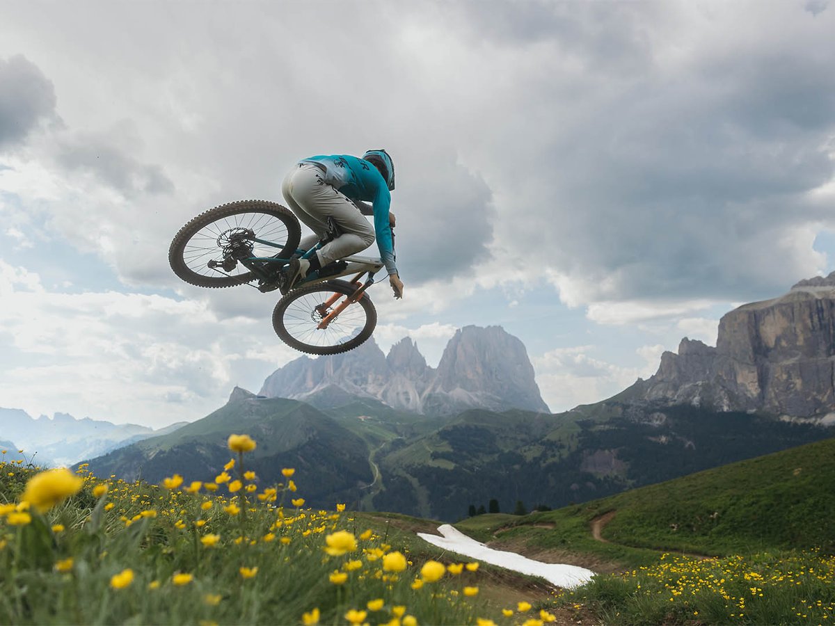 Mountain biker in teal Yeti kit catching air off a jump, bike angled nose-down, alpine peaks and yellow wildflowers below.