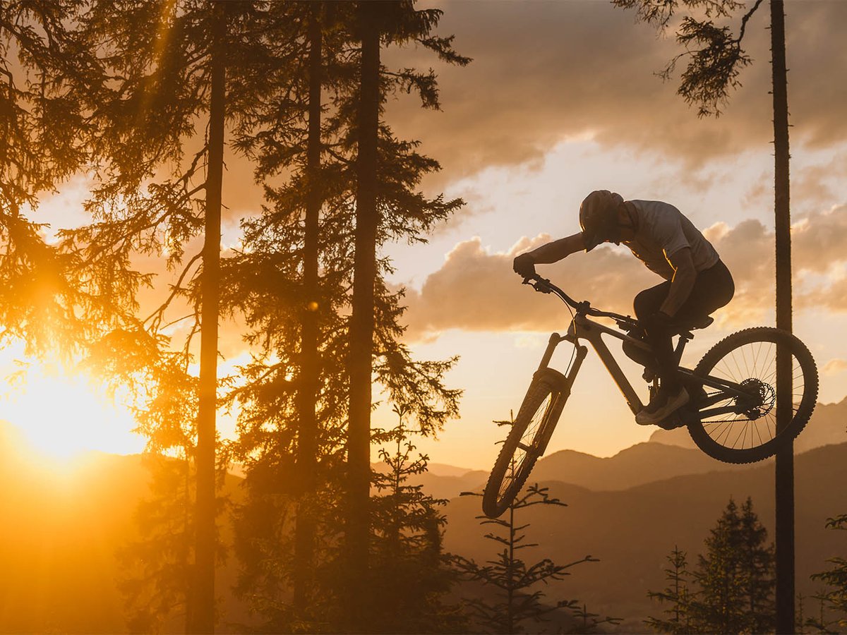 Silhouette of a mountain biker jumping a Yeti bike through a forest clearing at sunset, sun flaring low behind pine trees.