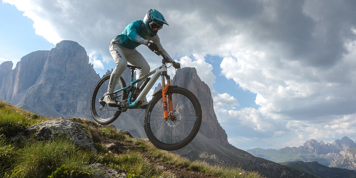 Mountain biker in teal Yeti kit descending a rocky alpine trail on a teal Yeti full-suspension bike with orange Fox fork, jagged Dolomite peaks in the background.