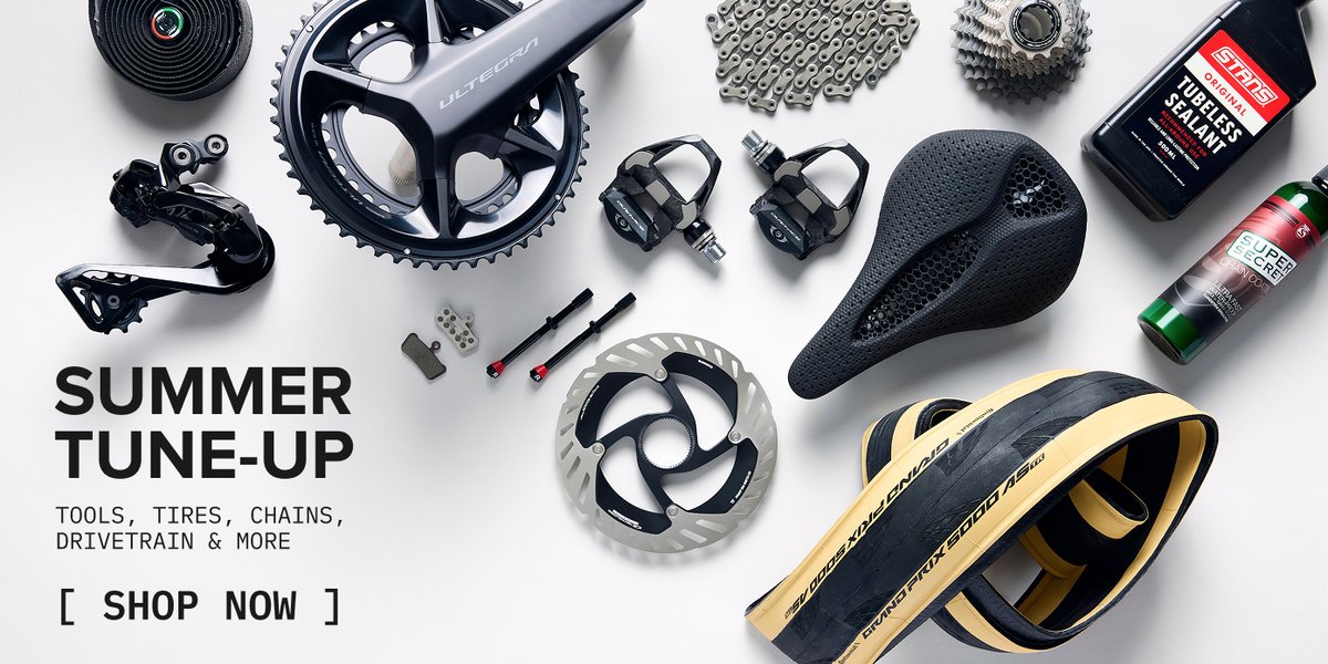 Flat-lay of road bike components — crankset, chain, cassette, pedals, saddle, brake rotor, tubeless sealant, and tire.