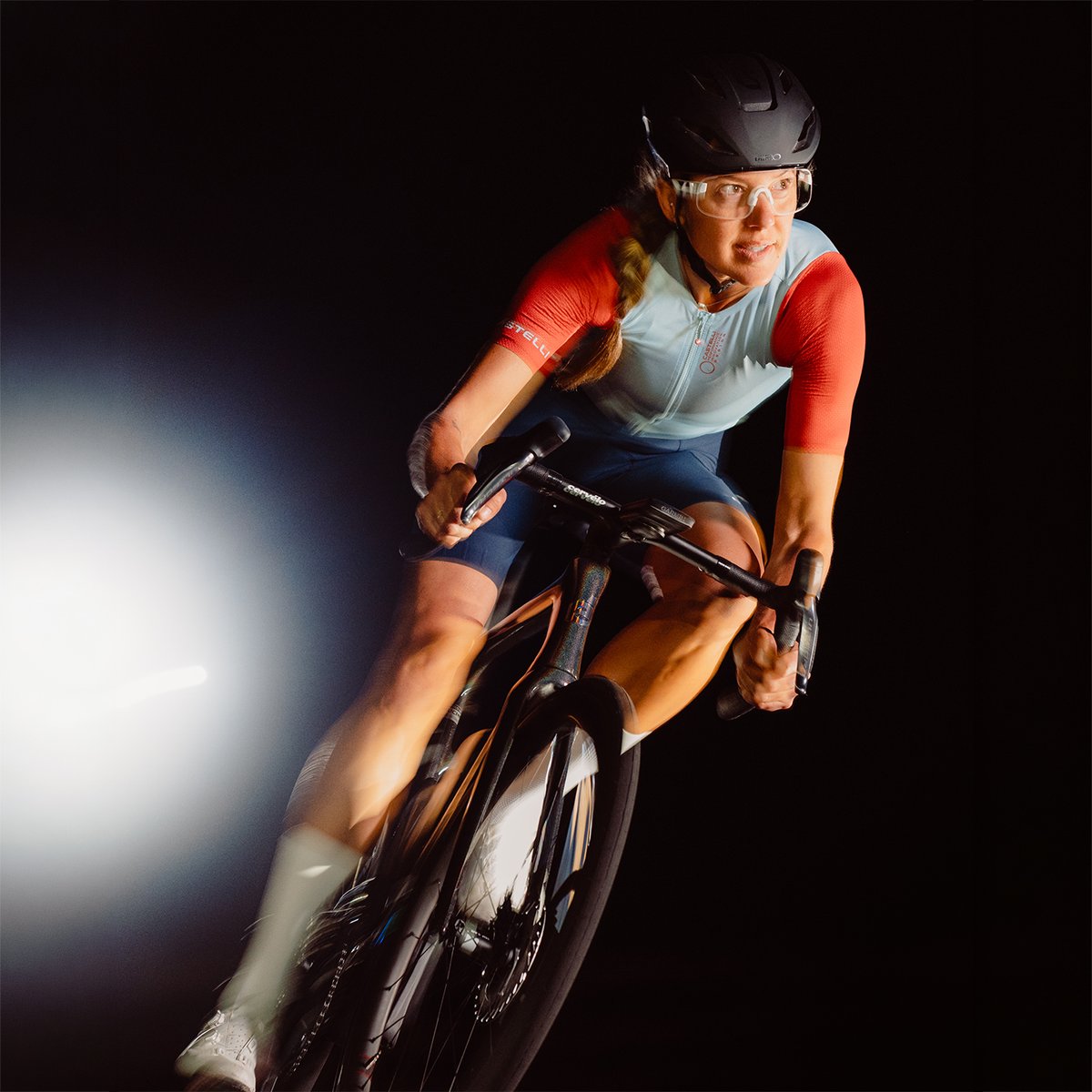 Female cyclist in a light blue and red Castelli kit riding a white Cervélo road bike toward the camera against a black background with a spotlight to the left.