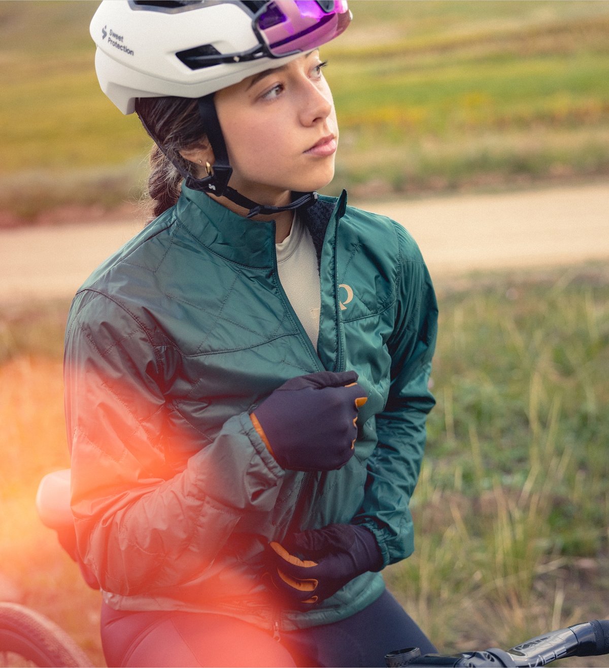 A cyclist wearing a white helmet, reflective sunglasses, teal-green jacket, and black gloves zips up her jacket.
