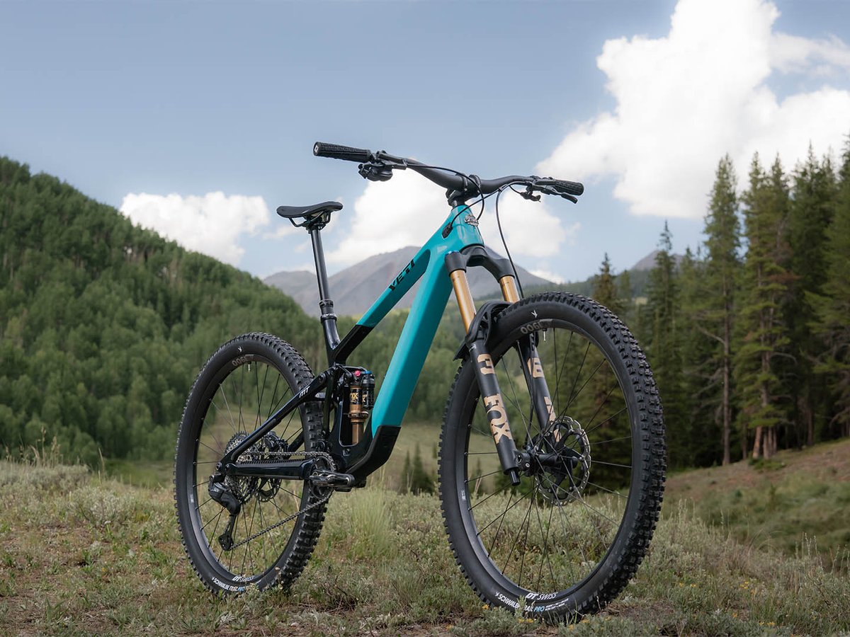 Full profile of the Yeti MT full-suspension mountain bike in turquoise, parked in an alpine meadow with conifers and peaks behind.