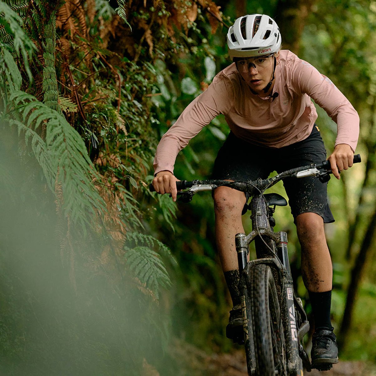 A woman mountain biker charges toward the camera on a mud-covered Santa Cruz Tallboy, navigating a tight forest trail lined with ferns.