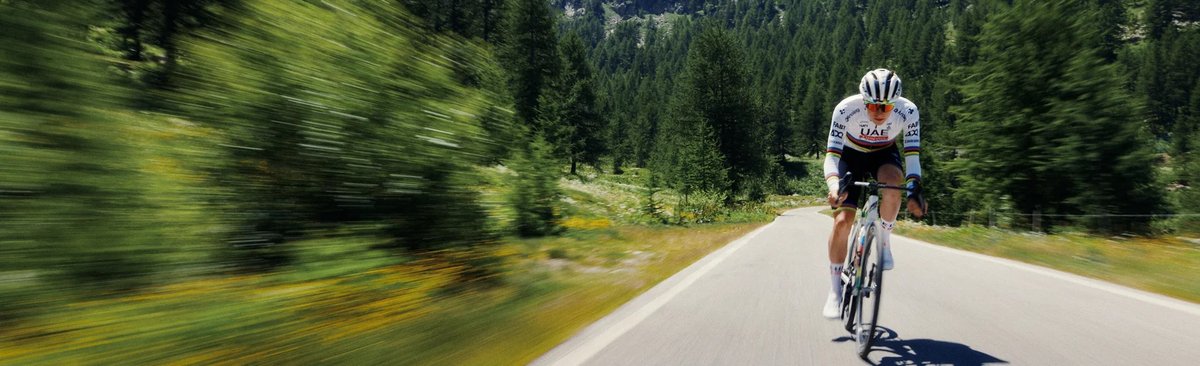 wide shot of cylict riding down paved road with forested mountain in the background.