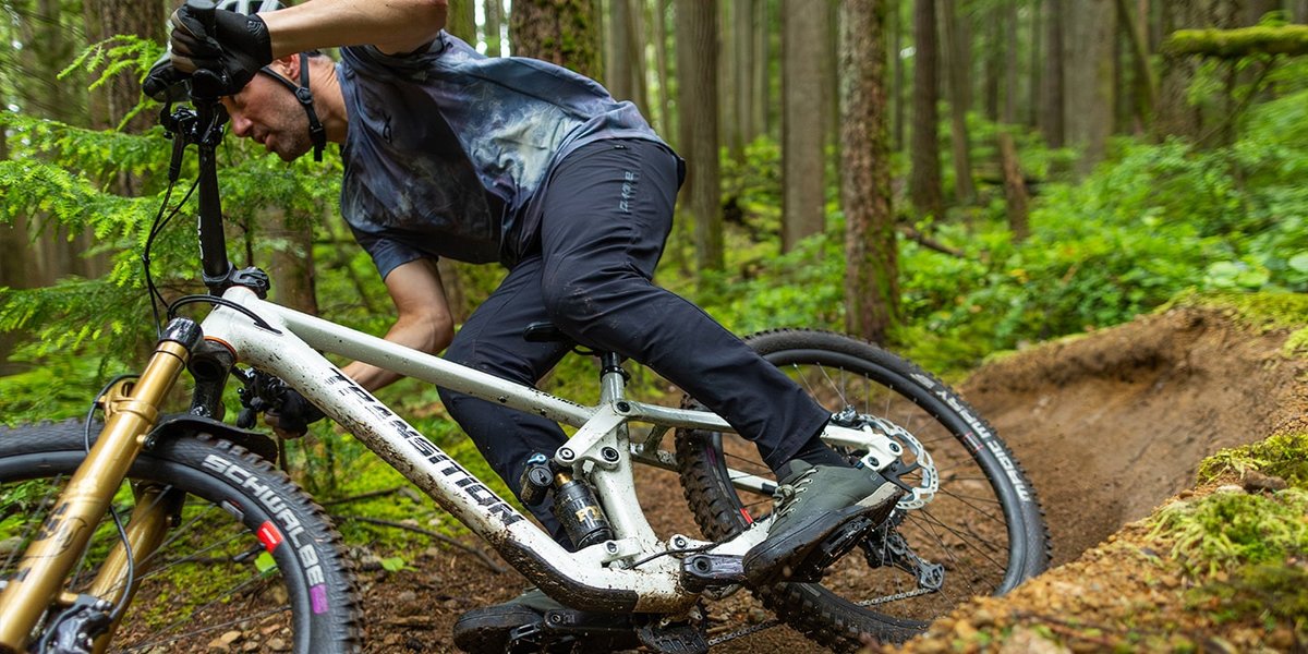 Rider in a gray jersey and black pants cornering hard on a mud-covered white Transition full-suspension mountain bike on a bermed dirt trail in a dense forest.