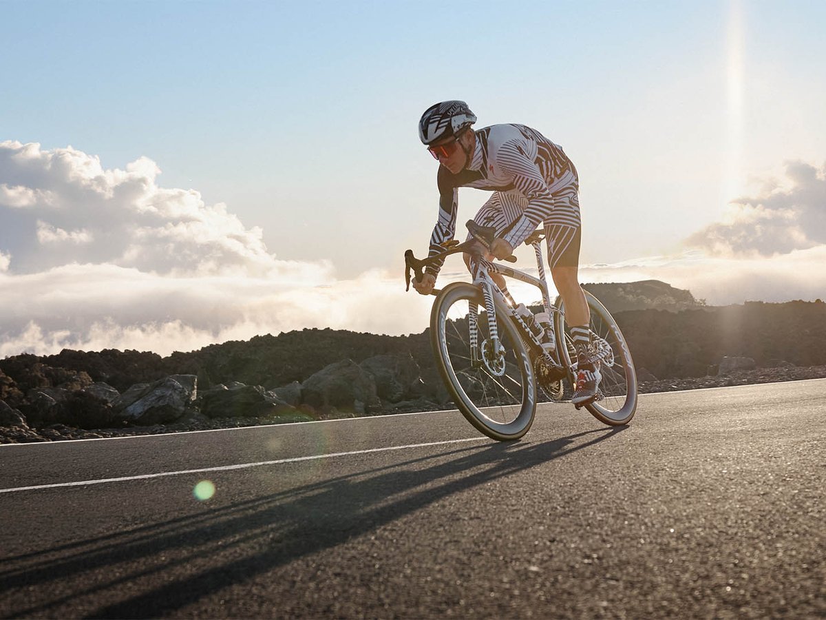 Road cyclist in a black and white graphic kit descending on a white S-Works Tarmac SL9 on a volcanic lava-field road, shot low against a backlit sky.