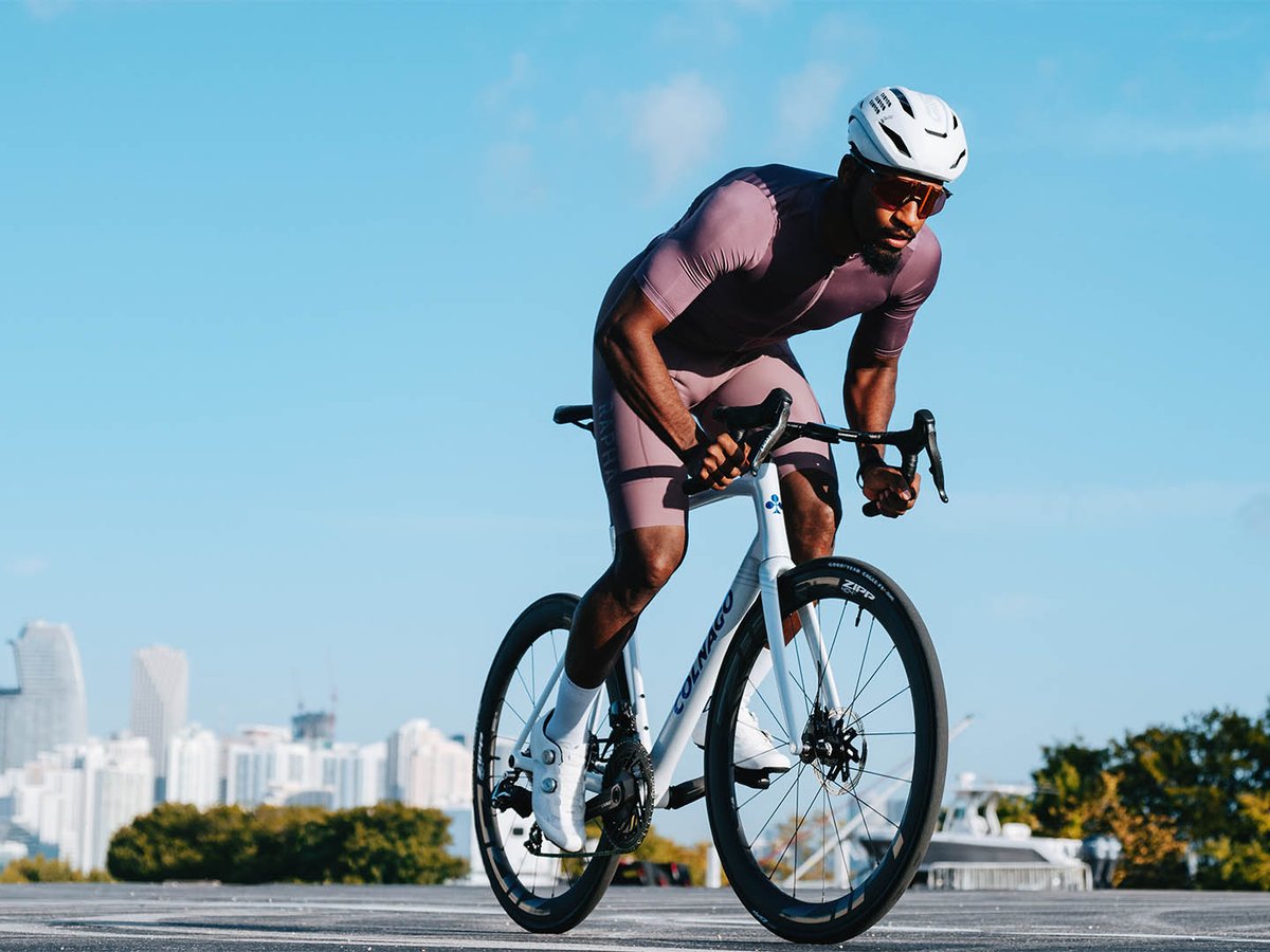 A male cyclist rides a white Colnago road bike fitted with Zipp 404 wheels in an aggressive position, with a city skyline visible in the background under a clear blue sky.