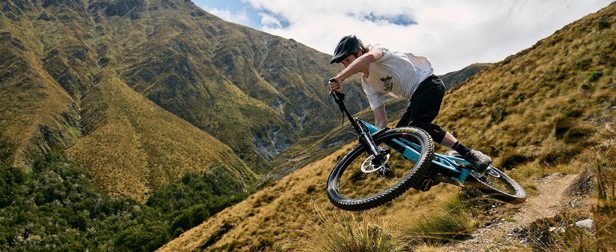 Mountain bike rider launching off steep alpine singletrack into the air above open mountain valley terrain on a sunny day