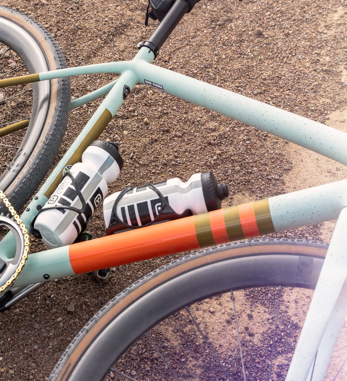 A close-up of a light blue and orange bicycle lying on gravel, featuring two water bottles mounted on the frame.