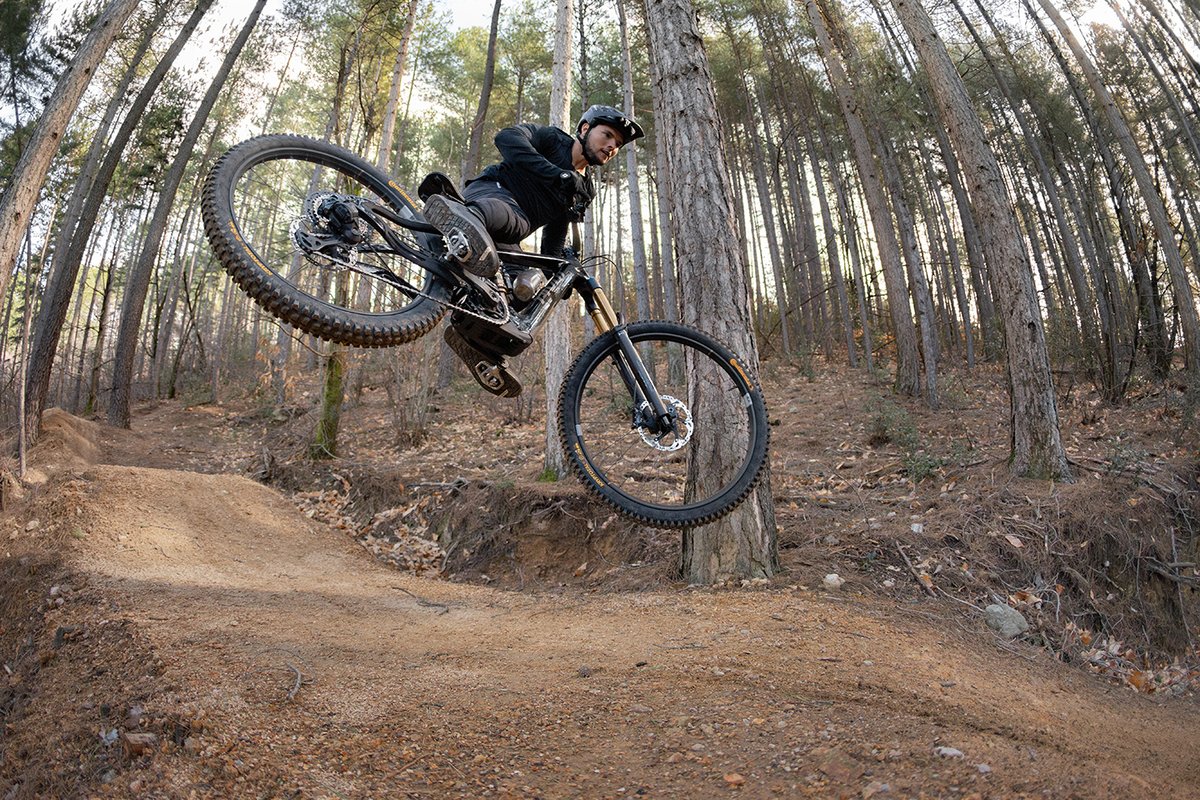 Rider whipping full-suspension eMTB off dirt jump wearing all-black MTB kit and helmet on dry forest trail in autumn conditions