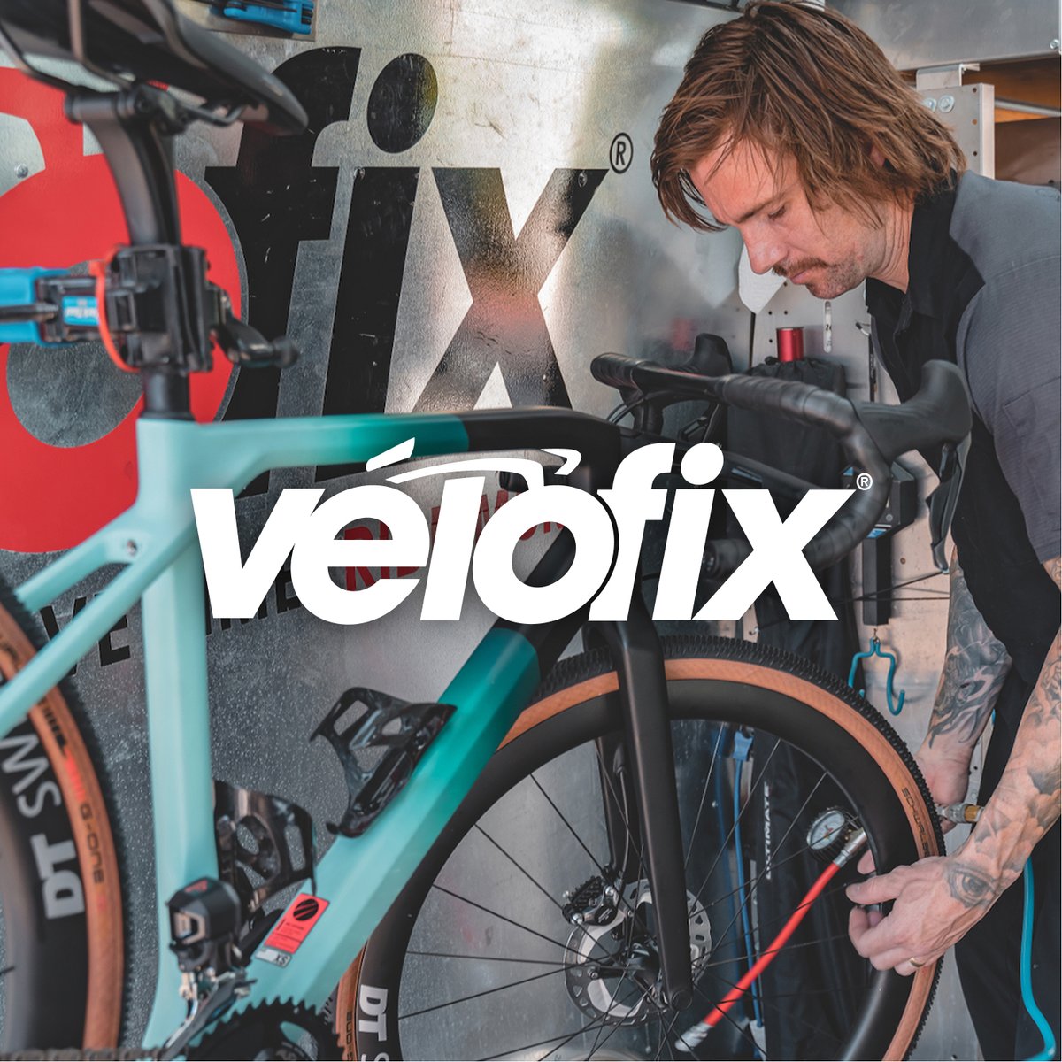 Mechanic inflating the front tire of a teal gravel bike mounted in a repair stand inside a workshop, featuring the Velofix logo.