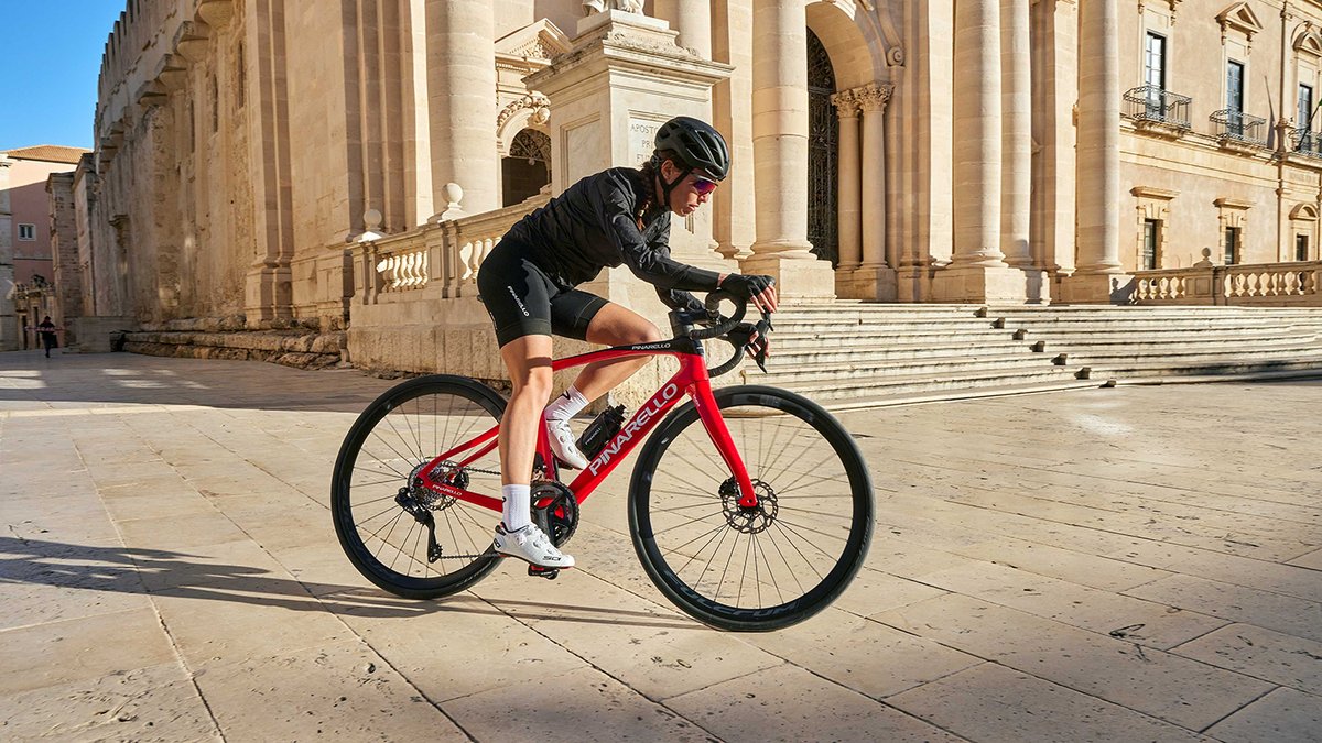 Road cyclist wearing cycling jersey, bib shorts, and road cycling shoes riding red Pinarello carbon road bike across historic European stone plaza