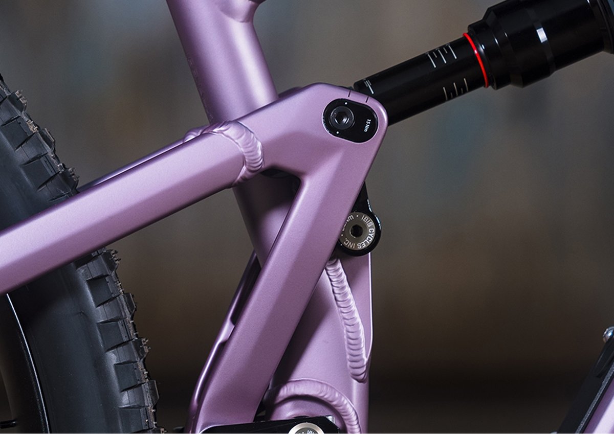 Close-up view of the rear suspension linkage on a matte rose full-suspension mountain bike. The image shows the intricate frame welds, pivot hardware, and part of the black rear shock above the rear wheel, highlighting the engineering and craftsmanship of the bike’s frame.