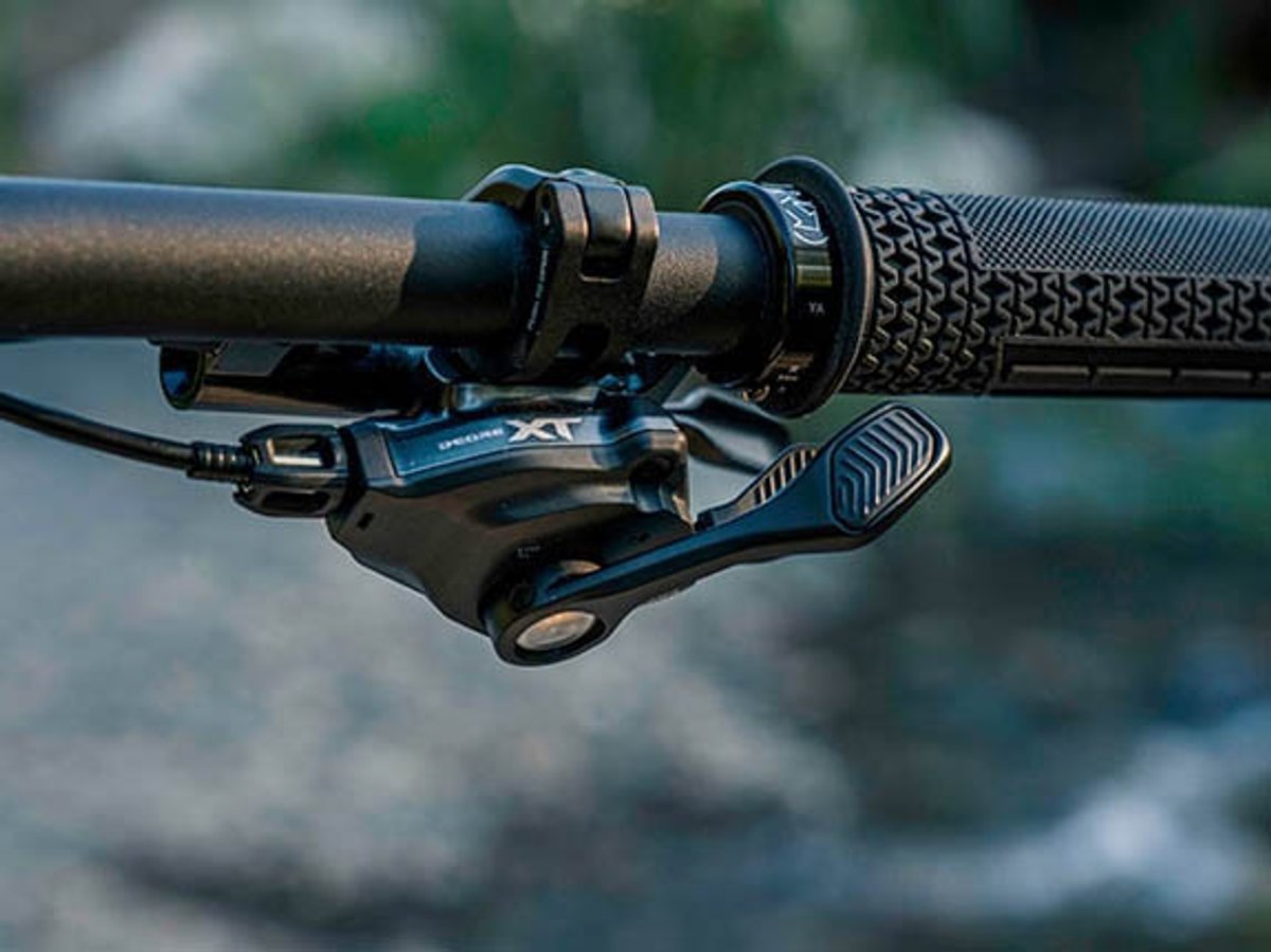 Shimano Deore XT shifter and brake lever on a handlebar, straight-on view showing the shift paddle detail, with blurred rocky background.