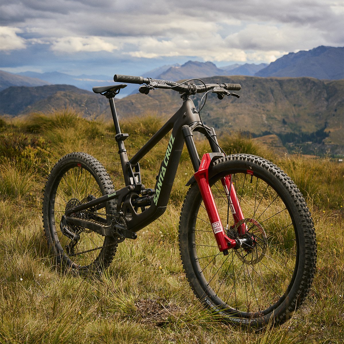 Sant Cruz nomad Bike standing upright in grass with mountain ridges in teh background.