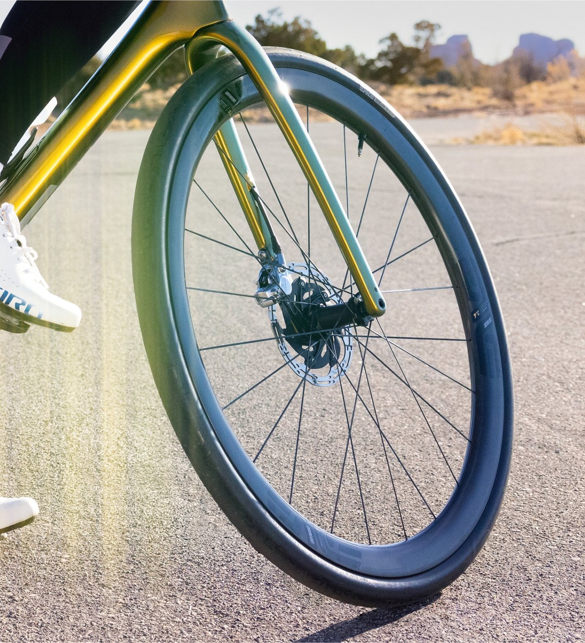 A close-up of the front wheel and fork of a road bike on pavement, with part of a cyclist’s white shoe visible and a dry, sunny landscape in the background.