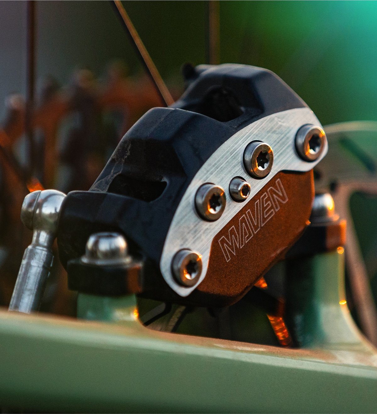 A close-up of a black and silver bicycle brake caliper with the word "MAVEN" engraved on it, mounted on a green bike frame.