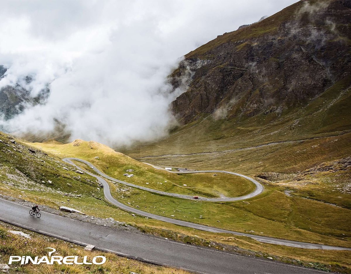 Cyclist riding down winding mountain road through grassy hills with cloudy valley in the background.
