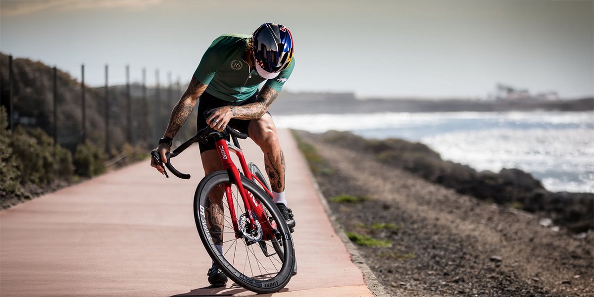 Heavily tattooed road cyclist in a green jersey and Red Bull helmet cornering aggressively on a red road bike  on a coastal path, ocean visible behind.