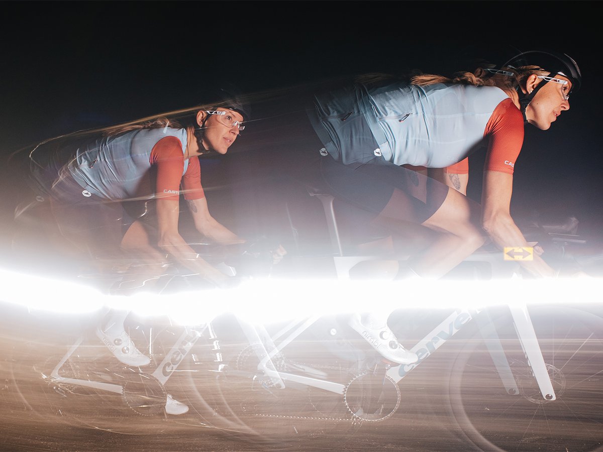 Multiple-exposure shot of a female cyclist in a light blue and red Castelli kit on a white Cervélo, with overlapping ghost images and a horizontal light streak across the frame.