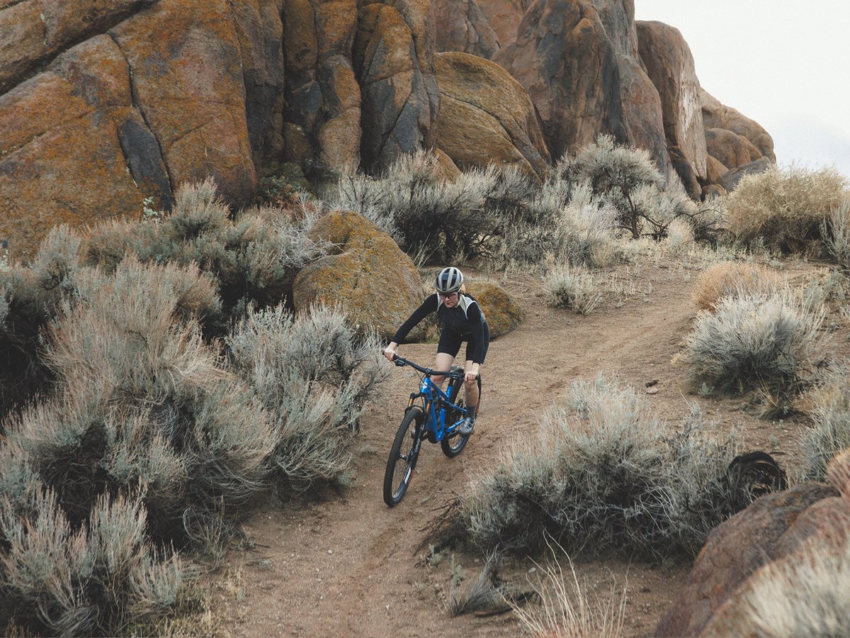 A mountain biker tears down a winding singletrack trail surrounded by dry shrubs and large orange-brown rock formations in a desert landscape.