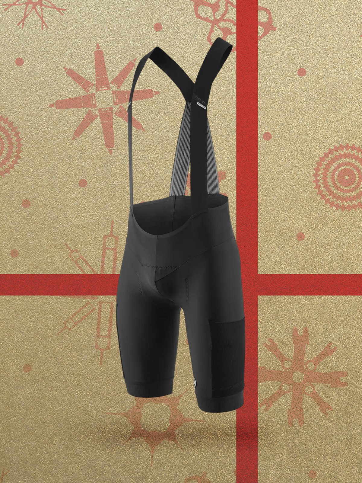 A pair of black cycling bib shorts with shoulder straps shown against a gold background decorated with red holiday patterns and red stripes.