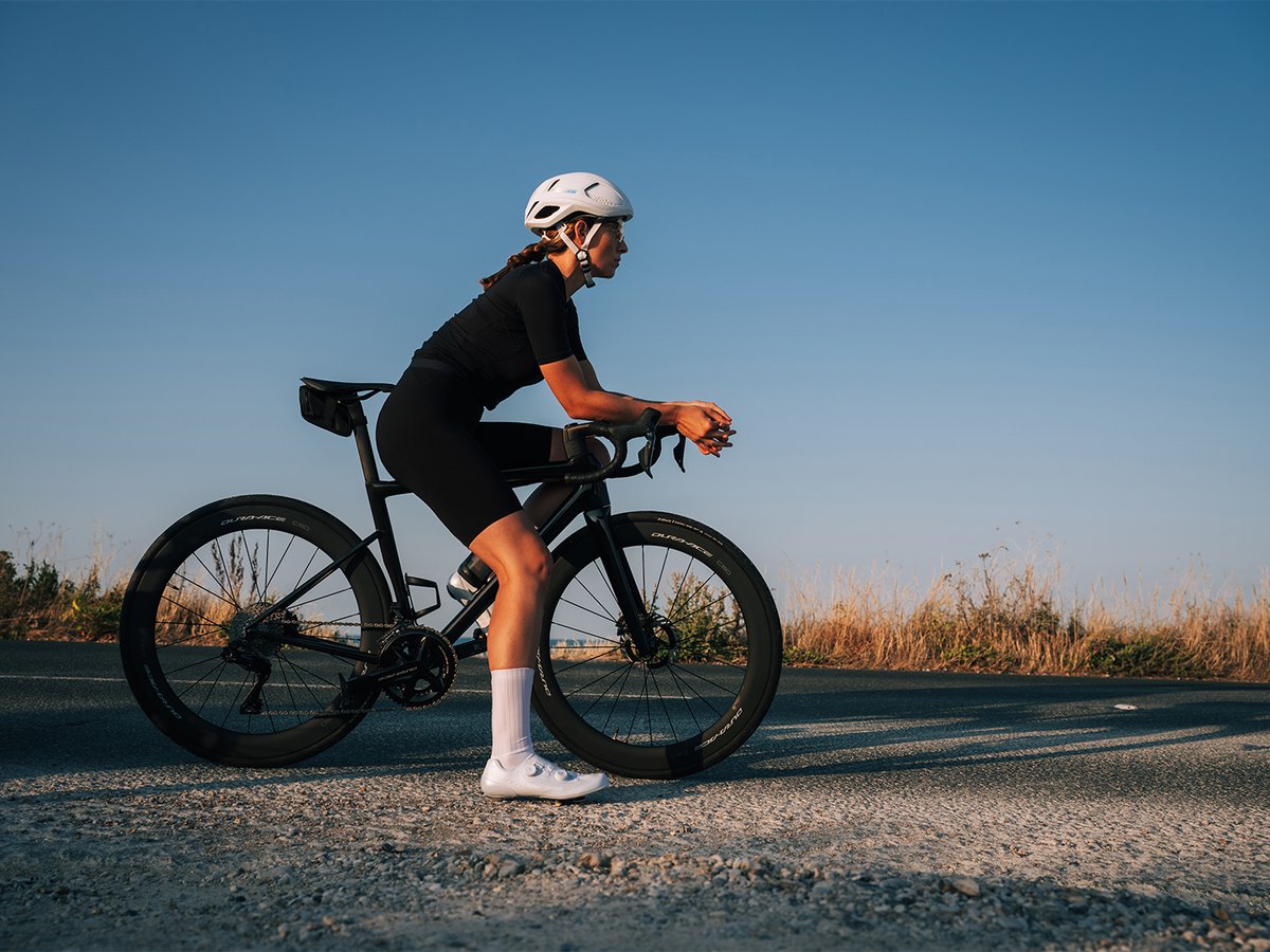 Cyclist in black kit standing beside a road bike, profile view, white S-PHYRE shoe visible, Dura-Ace wheel legible, clear sky background
