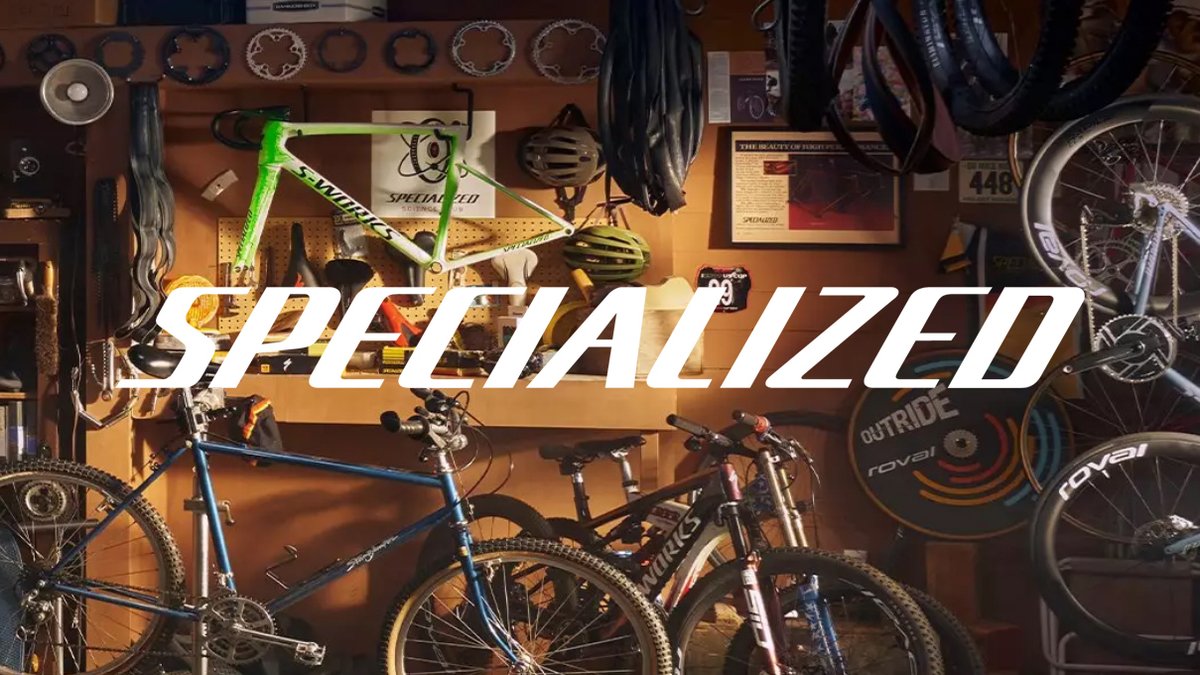 A cluttered, sun-drenched bicycle workshop filled with mountain bikes, hanging tires, and specialized tools, with the Specialized logo boldly centered in the middle of the frame.