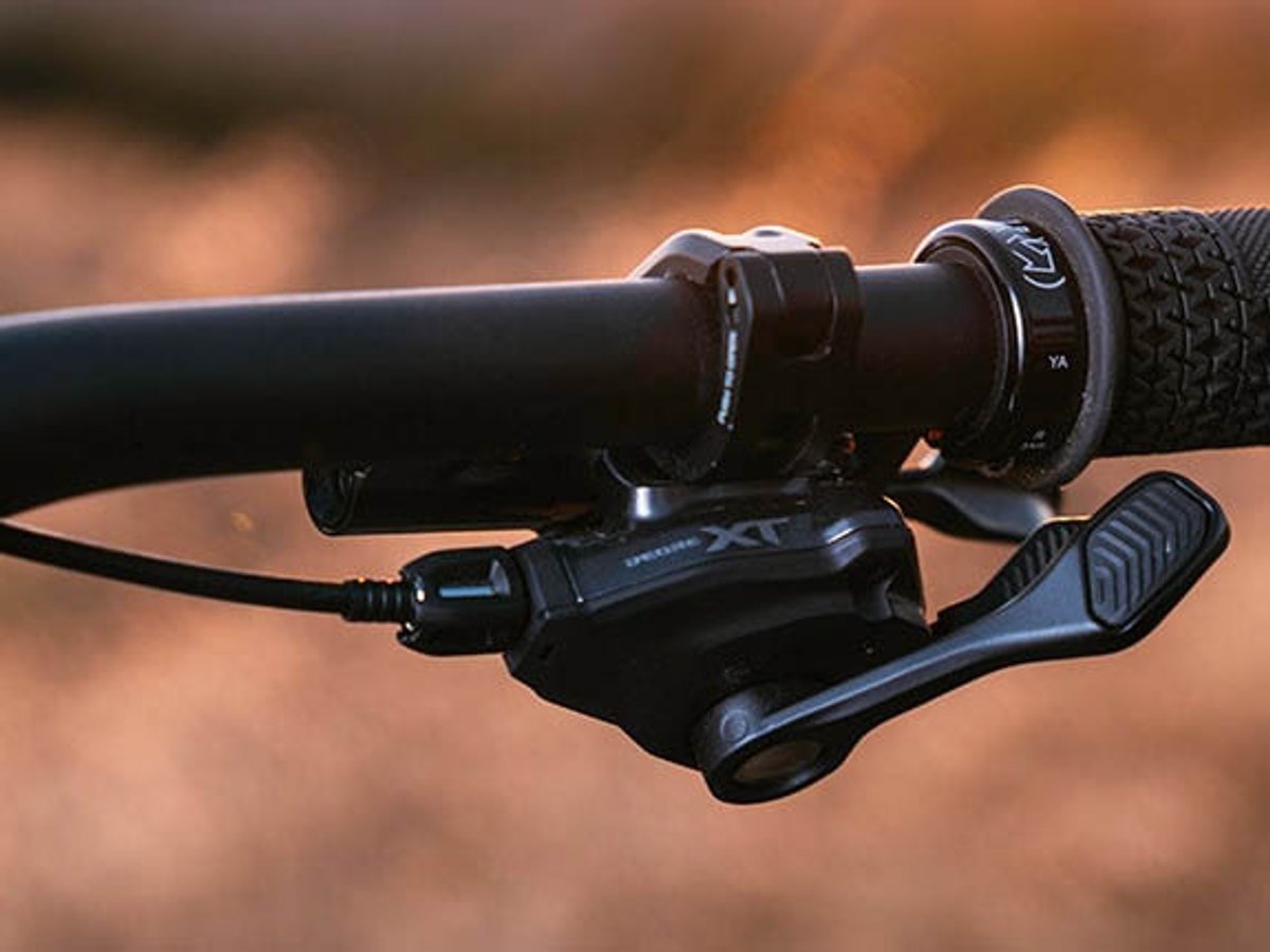 Shimano Deore XT shifter and brake lever on a handlebar, angled view showing the shift paddle, with warm blurred background.