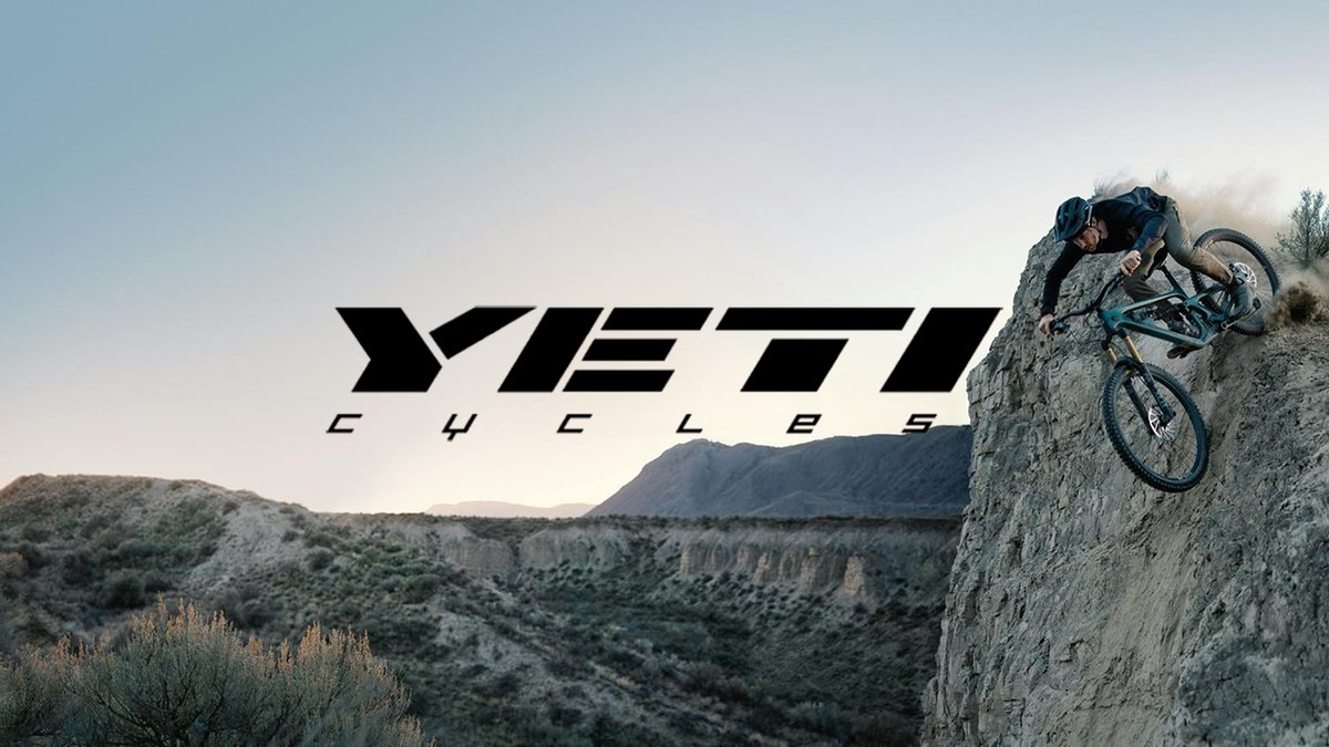 Mountain biker riding down a steep sandstone cliff edge, dust kicking up behind, with the "Yeti Cycles" wordmark overlaid on an arid canyon landscape.