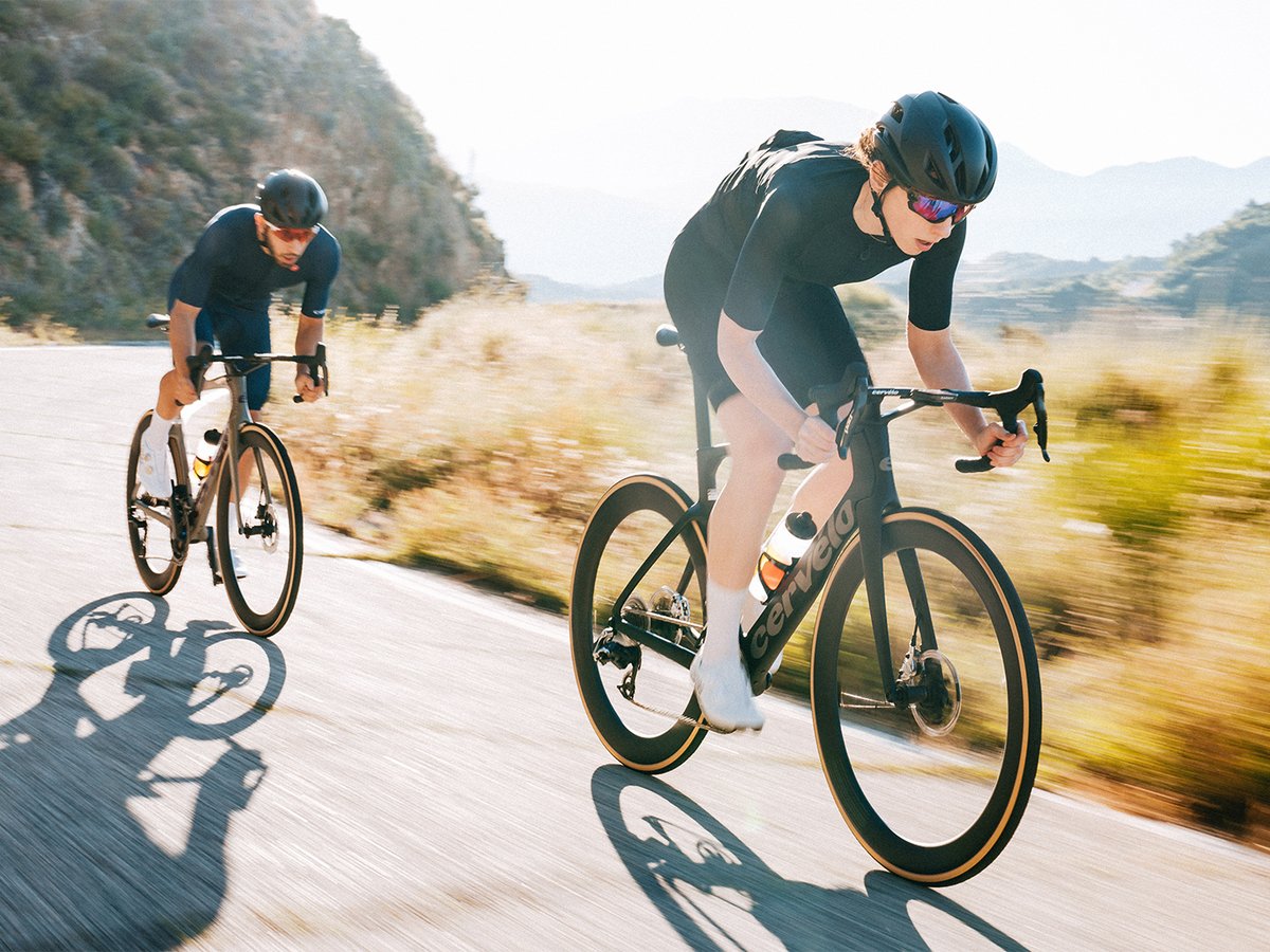 Two cyclists riding road bikes on a paved road in a dry, open landscape.
