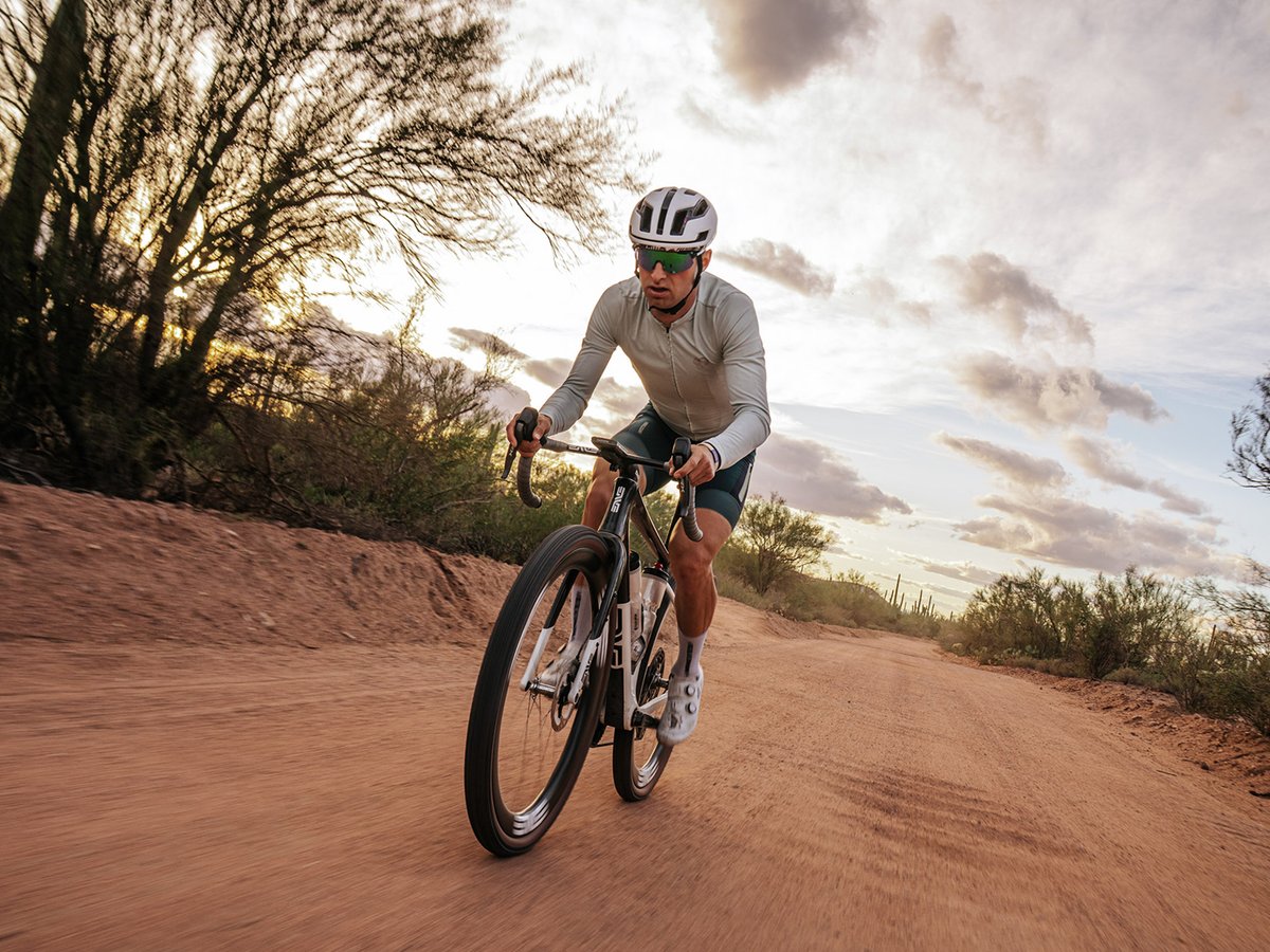 A male cyclist rides an ENVE gravel bike on a red dirt road through desert scrub at dusk.