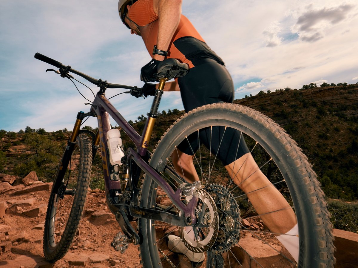 A rider climbs rocky desert singletrack on a purple full-suspension mountain bike with gold forks, drivetrain and rear wheel filling the frame.