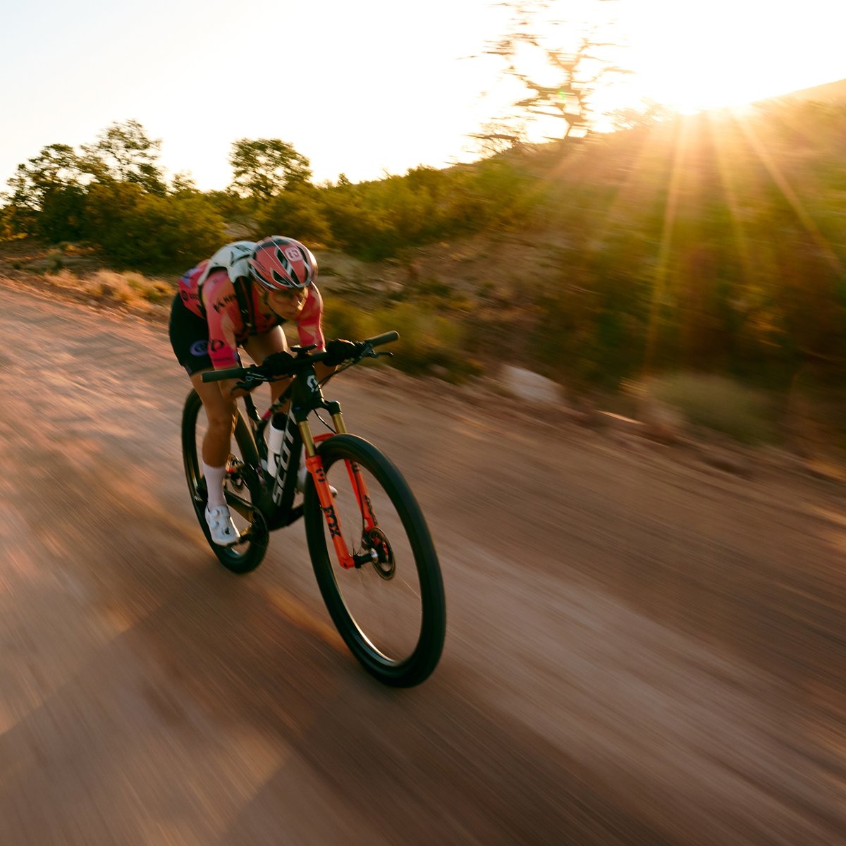 Hannah tucks on her mountain bike, speeding down a dirt trail, with the sun setting and casting rays through trees in the background.