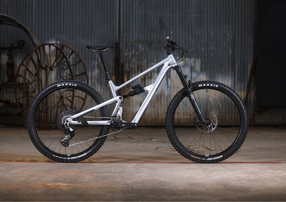 A silver full-suspension mountain bike with Maxxis tires stands on a concrete floor in front of a corrugated metal wall.