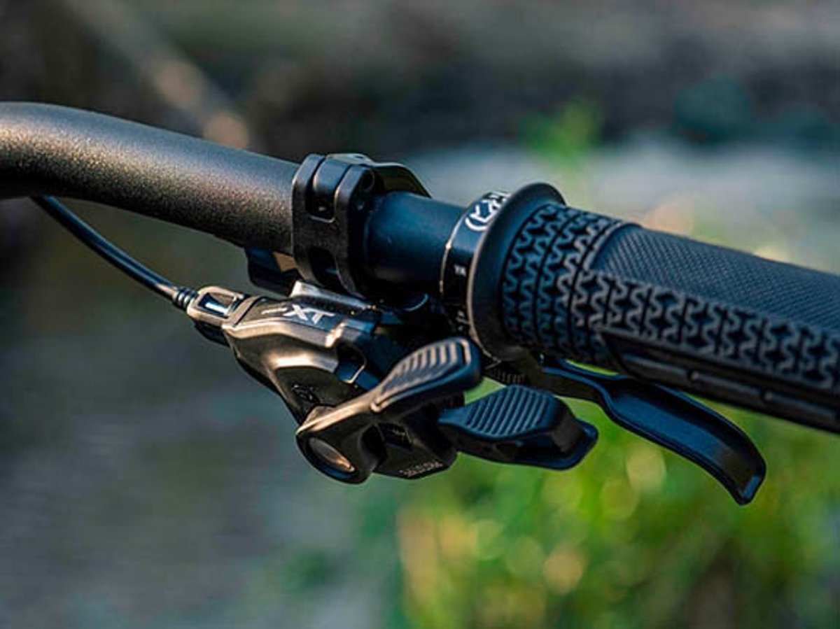 Shimano Deore XT shifter and brake lever mounted on a mountain bike handlebar, shot close up with blurred greenery behind.