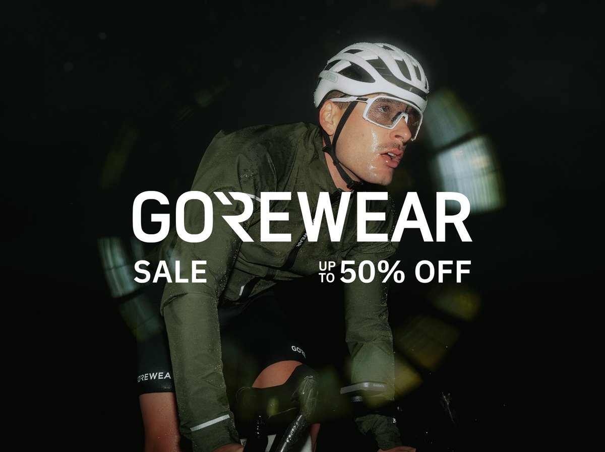 A cyclist wearing a white helmet and a dark green GOREWEAR jacket rides in  nighttime conditions, with water droplets visible on the jacket and blurred lights in the dark background. Overlaid is bold text reading “GOREWEAR Sale up to 50% off shop now” 