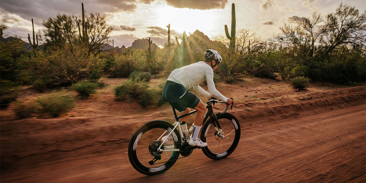 A cyclist rides an ENVE gravel bike at speed along a red dirt road through a desert landscape at golden hour, with saguaro cacti and mountains visible in the background.