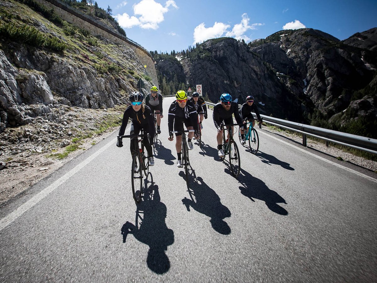 Group of five road cyclists climbing a narrow alpine road from a low camera angle, with rocky canyon walls and a blue sky above.a