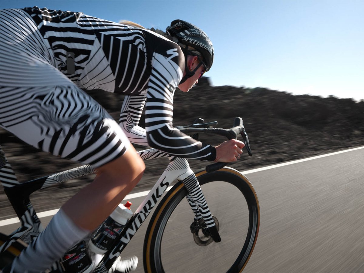 Panned side shot of a cyclist in a black and white Specialized kit sprinting on a white S-Works Tarmac SL9 with tan-sidewall wheels, lava fields blurred behind.