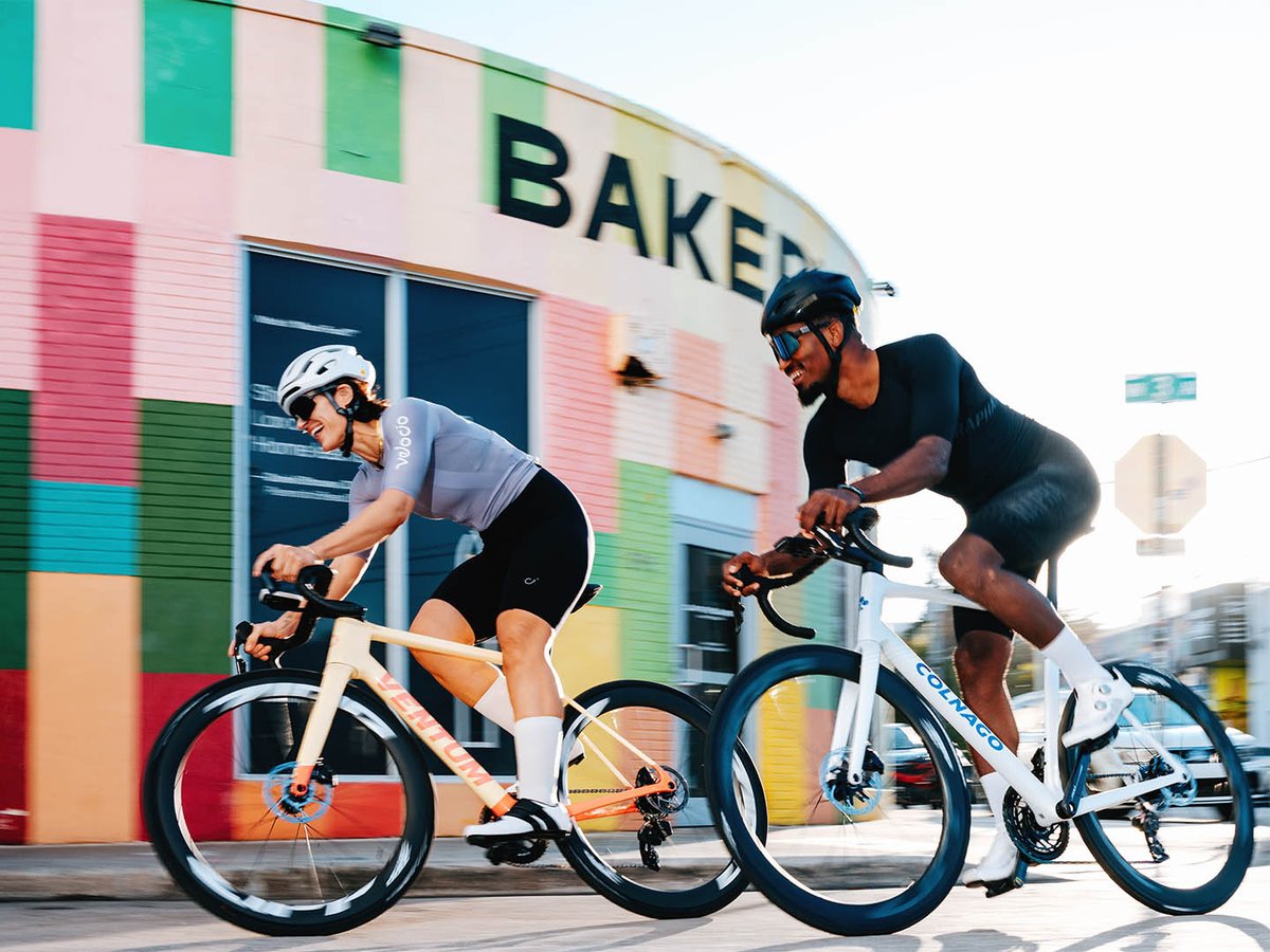Two cyclists ride road bikes with Zipp 404 wheels past a colorful building in an urban setting, one on a Ventum and one on a Colnago.