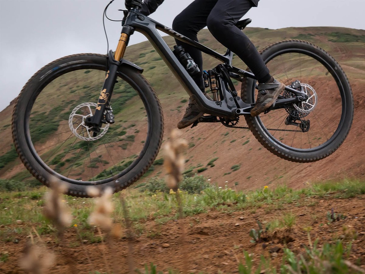 Low-angle detail of a carbon Yeti full-suspension mountain bike with Fox fork mid-pedal on a red-dirt hillside trail.
